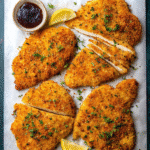 Golden-brown baked chicken cutlets arranged on parchment paper with fresh herbs, lemon wedges, and a small bowl of dark sauce